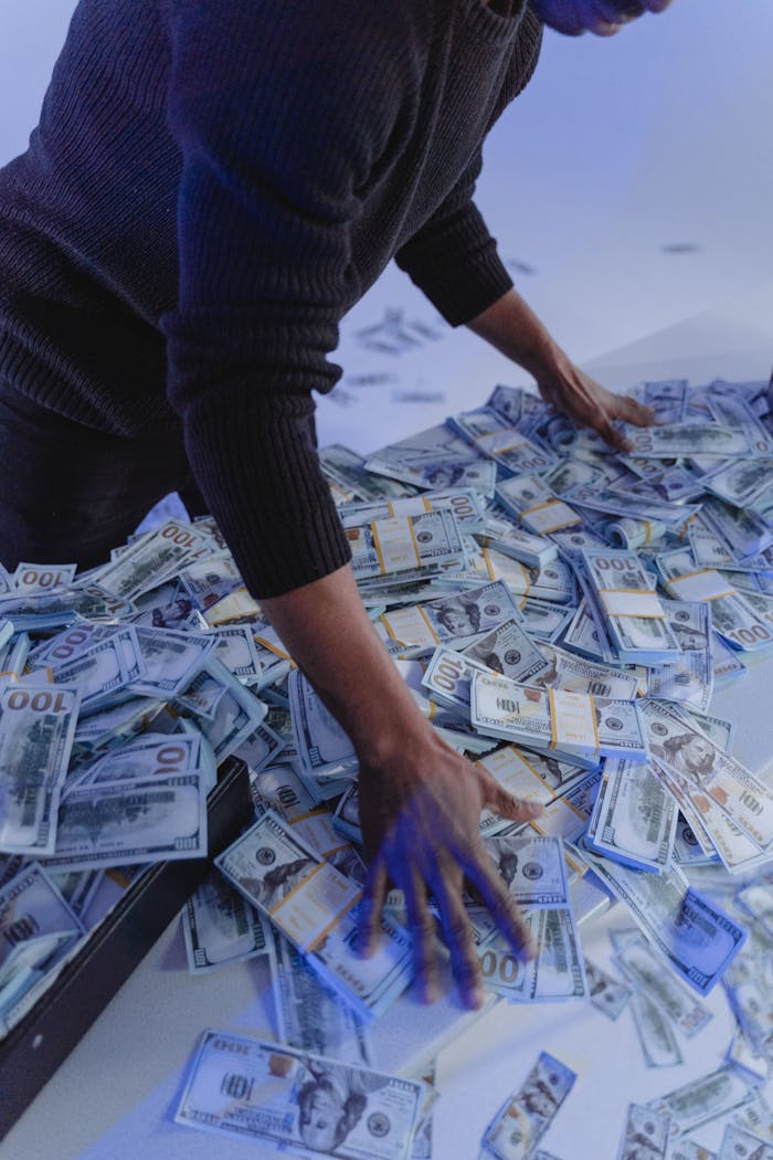 Adult man organizing a massive pile of US dollar bills indoors in dynamic lighting.